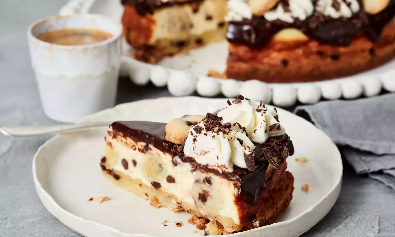 Ein Stück Cookie Dough Cheesecake mit Schokoglasur, Sahnetupfen, Keksstückchen und Schokoraspeln auf einem Teller neben einer Tasse Espresso