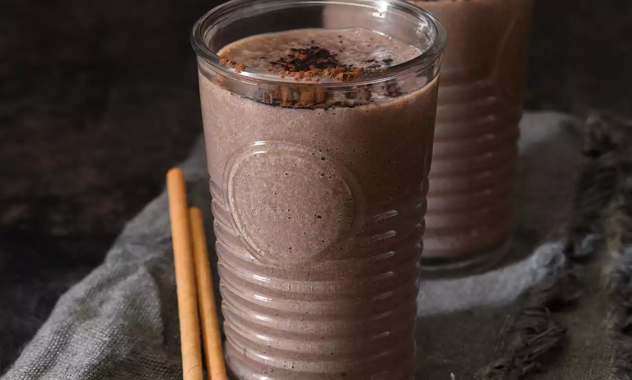 Hnědé smoothie jemné textury ve vysoké sklenici s reliéfem, posypané tmavým práškem, vedle plátěného ubrousku a dvou tyčinek
