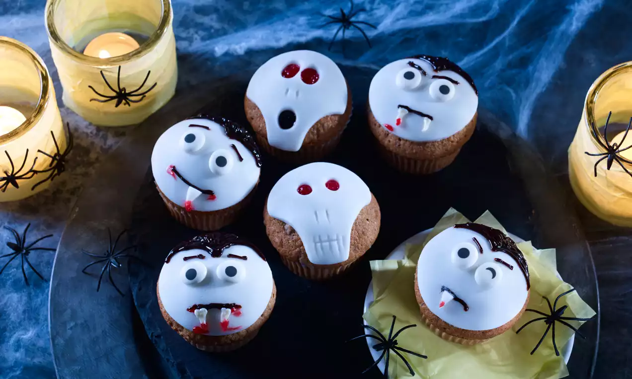 Muffins mit weißem Zuckerguss, verziert als Vampire mit roten Augen und Totenkopfgesicht, auf schwarzem Teller mit Spinnennetzdekor