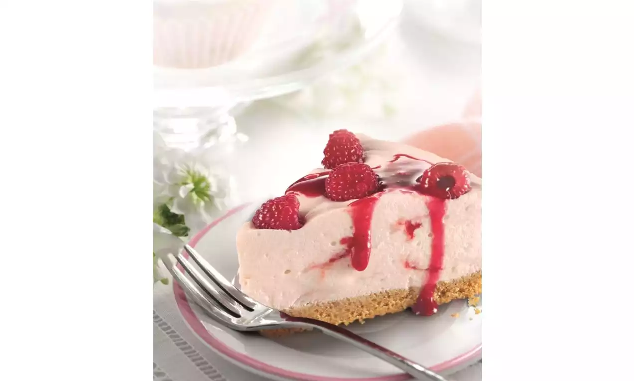 A wedge of pale pink raspberry cheesecake on a crisp biscuit base, topped with glossy red sauce and fresh raspberries on a white plate