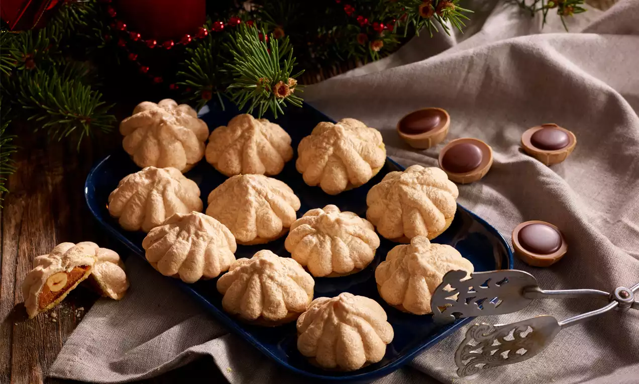 Goldbraune Toffifee-Kekse mit weicher, strukturierter Oberfläche in Blütenform auf einer dunkelblauen Platte, dekoriert mit Tannenzweigen