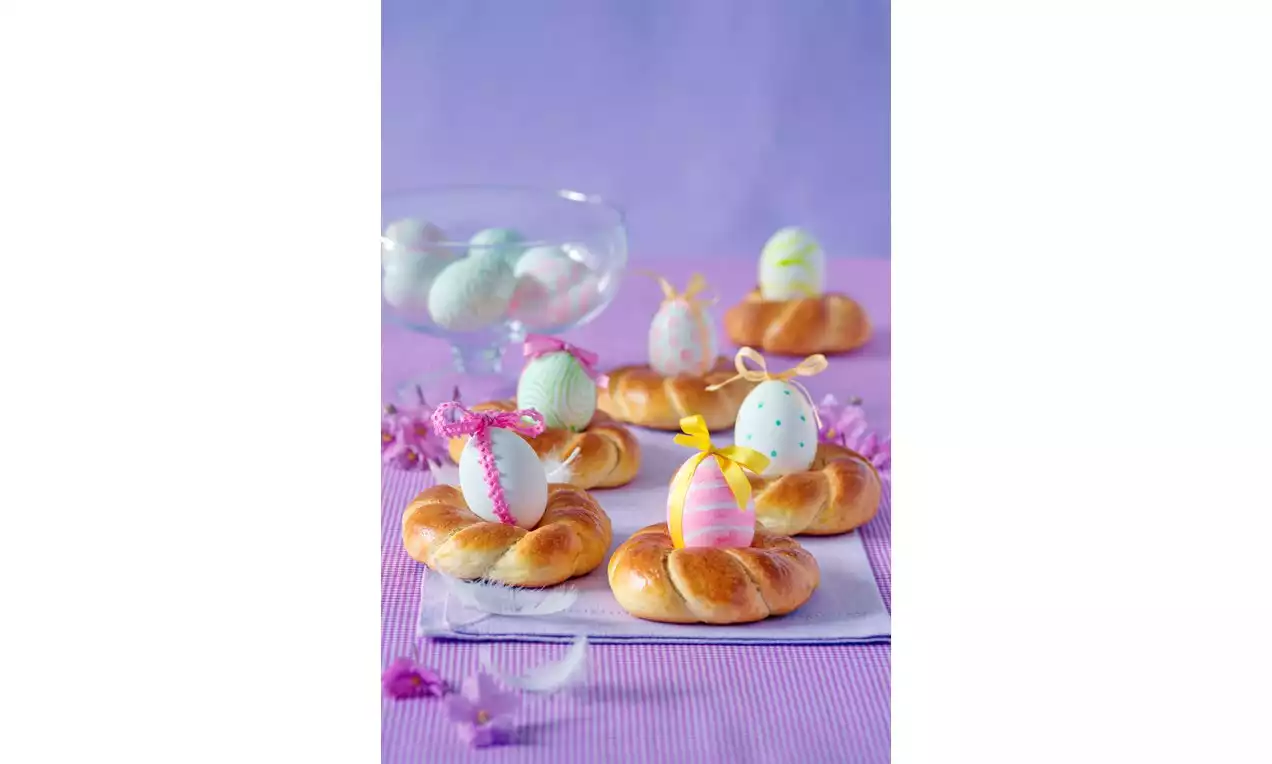 Zlatavé pletené věnečky s lesklými poleženými barevnými vajíčky na fialovém ubrousku, obklopené drobnými dekoracemi