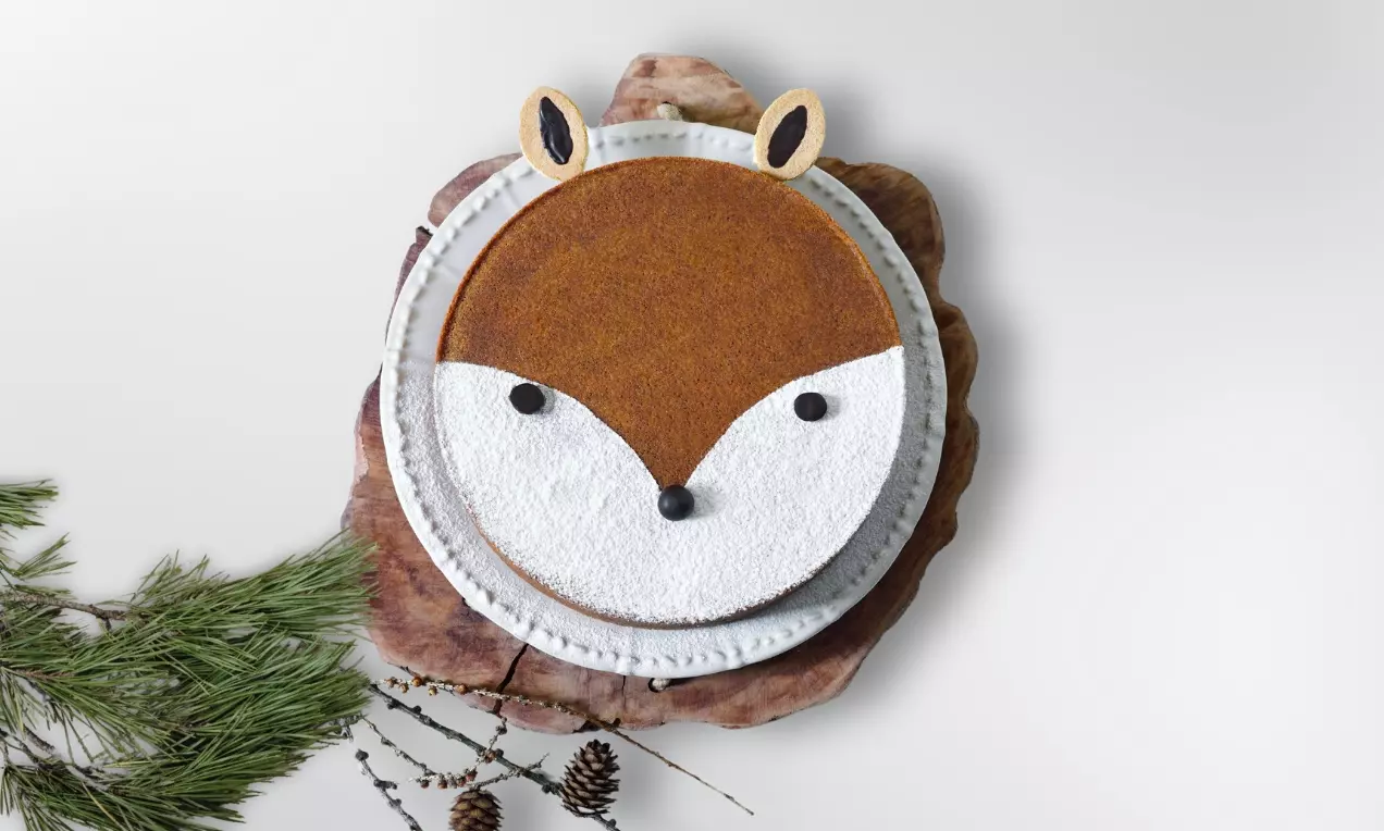 Mrkvový koláč ve tvaru liščí hlavy s hladkou oranžovou polevou, bílou cukrovou maskou, černými detaily na dřevěném podnosu vedle větviček a šišek