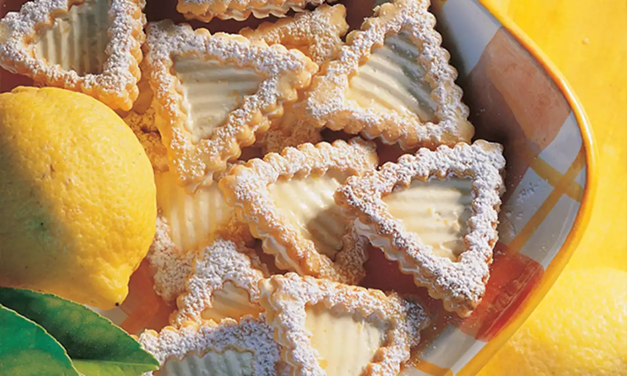 Driehoekige citroenkoekjes met geribbelde randen, licht bestoven met poedersuiker, naast een gele citroen in een kleurrijke schaal