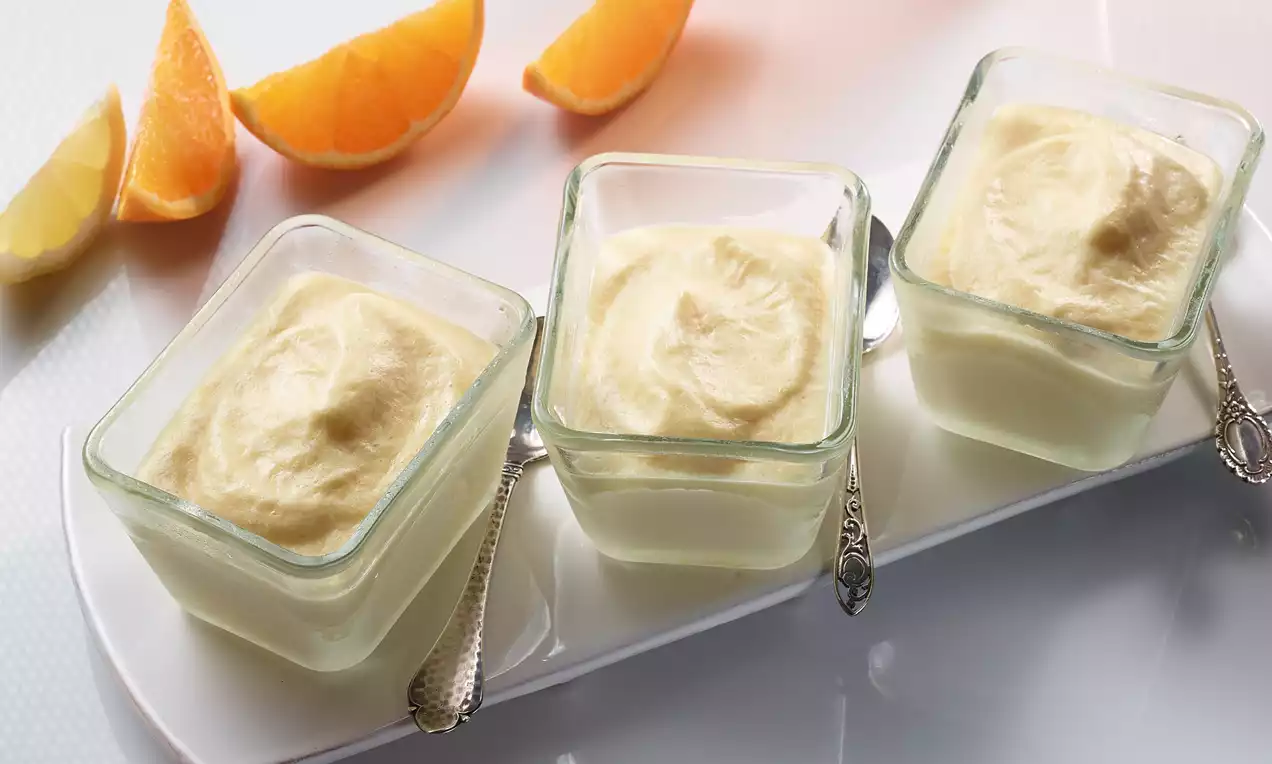 Drie vierkante glazen schaaltjes gevuld met romige, lichtgele mousse, geplaatst op een wit dienblad met zilveren lepeltjes en sinaasappelpartjes ernaast