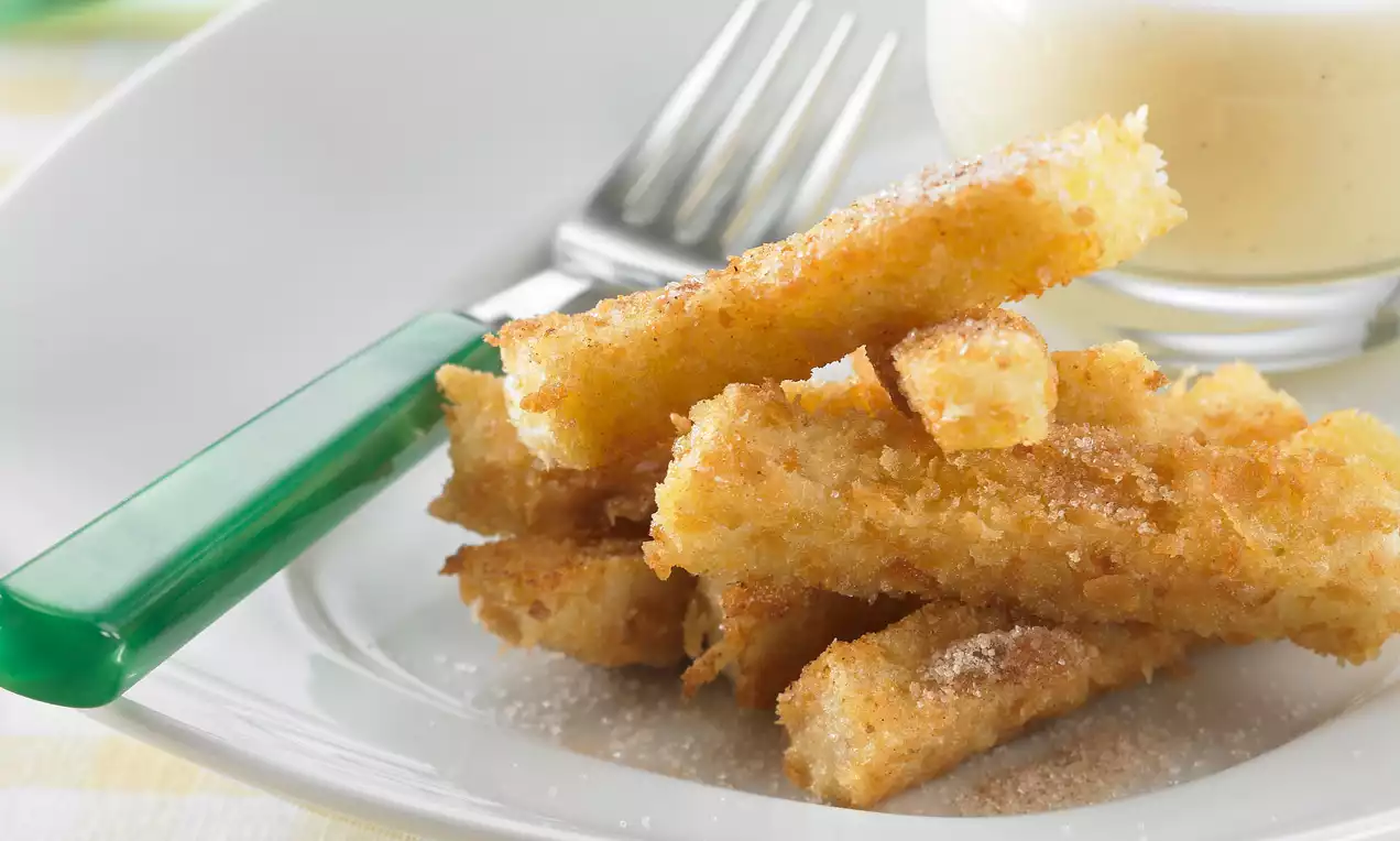 Stick dorati di pane croccante ricoperti di zucchero e cannella, disposti su un piatto bianco accanto a una ciotolina di crema chiara