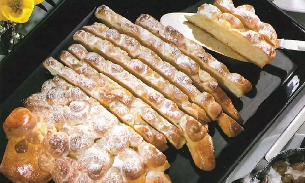 Pain doré en forme d'agneau stylisé, saupoudré de sucre glace, disposé en segments sur un plateau noir avec une fleur jaune en décoration