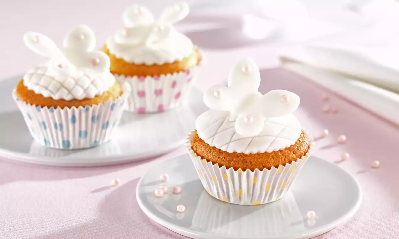 Goldene Muffins im gepunkteten Papierförmchen, gekrönt von weißem Fondant mit zarten Schmetterlingen auf rosa Tischdecke mit Perlen