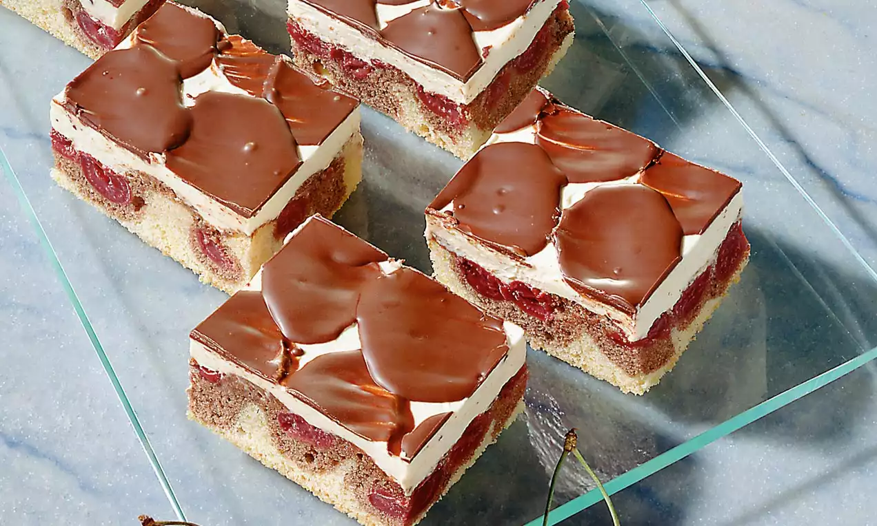 Pravokutni kolači s bijelom kremom, crvenim višnjama i mramornim čokoladnim preljevom, složeni na staklenom tanjuru