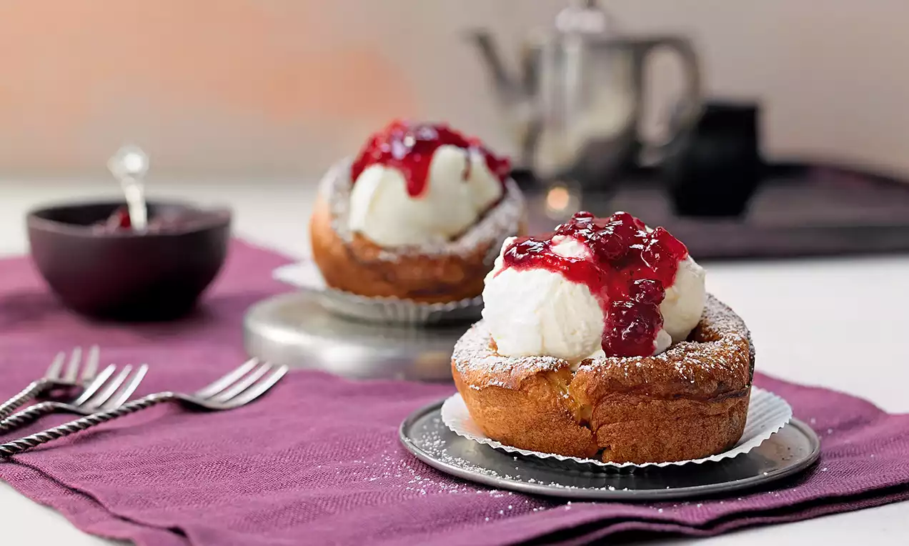 Goldbraune, fluffige Pfannkuchen-Muffins in Papierförmchen mit einer weißen Sahnehaube und glänzender roter Preiselbeersoße serviert