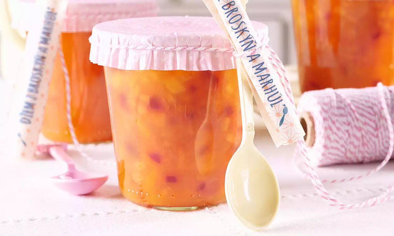 Sklenený pohár s oranžovým broskyňovo-marhuľovým džemom plný viditeľných kúskov ovocia, zakrytý jemným ružovým vrchnákom z papiera