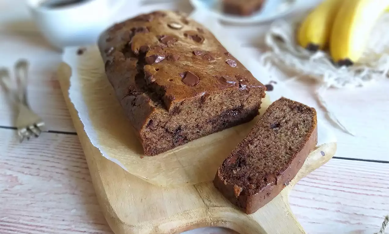 Banánový chlebík s hnedým povrchom, kúskami čokolády a orechov, položený na drevenej doske s jedným narezaným plátkom vedľa neho