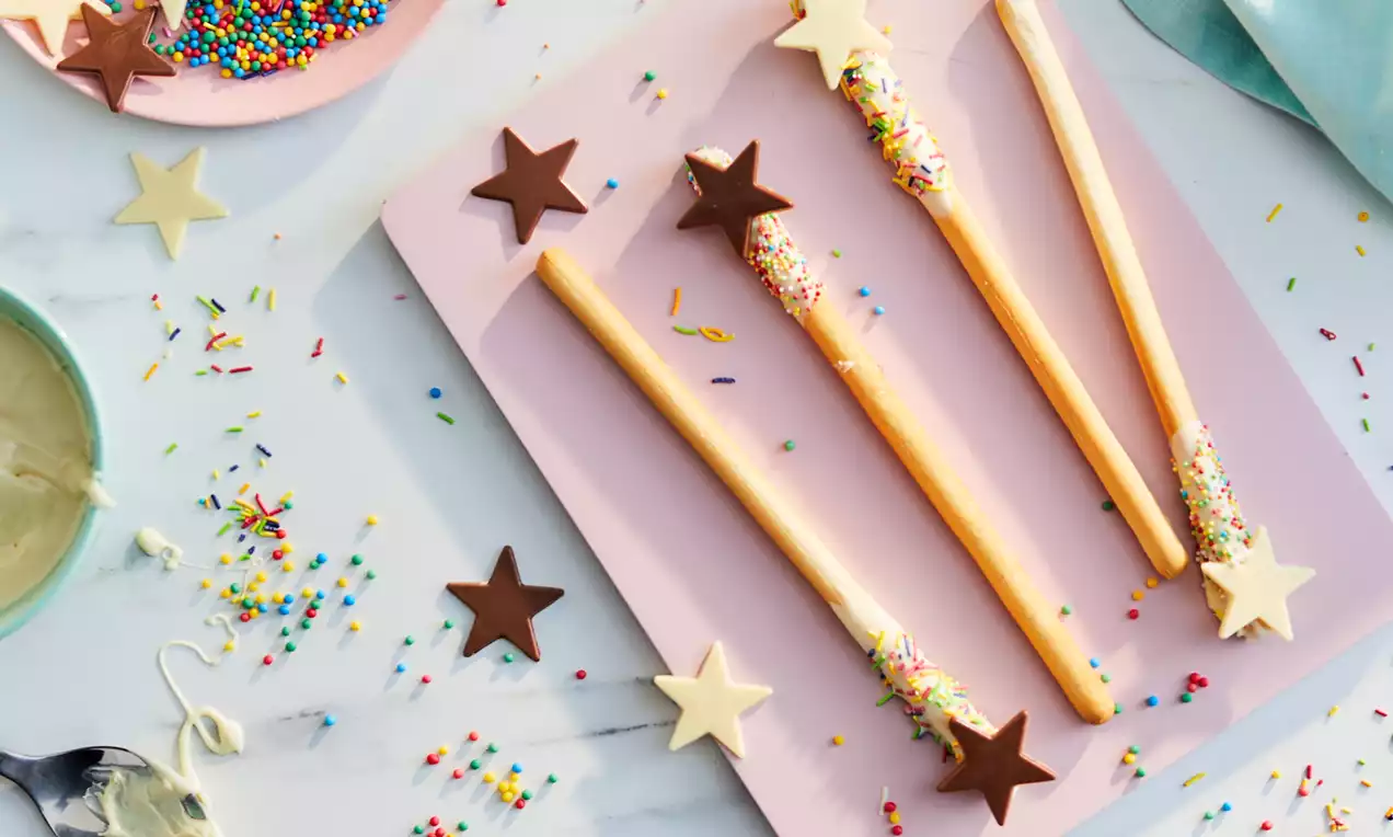 Golden biscuit wands topped with chocolate stars and sprinkled with multicoloured sugar on a pale pink tray amidst scattered decorations
