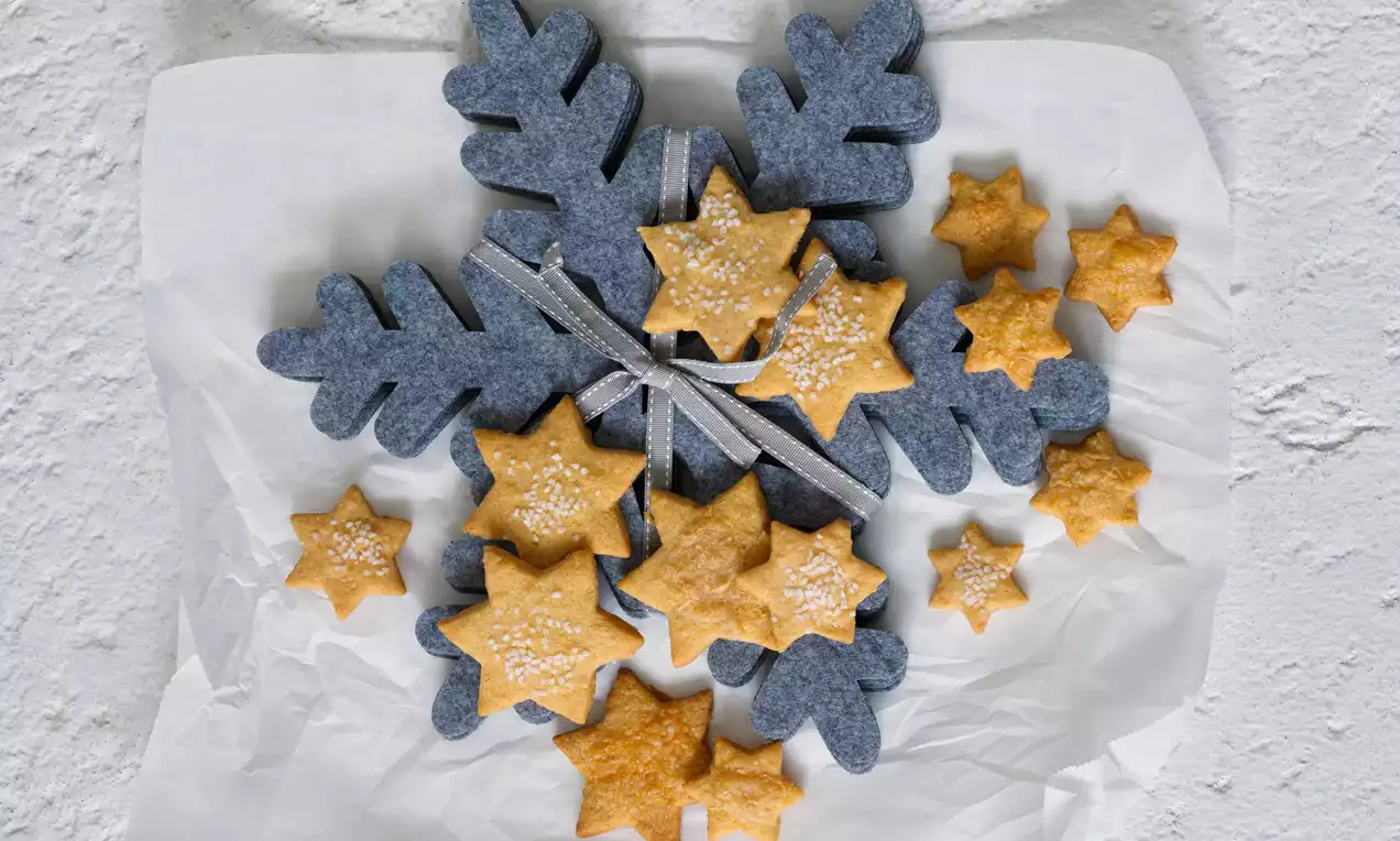 Sádlové cukroví v tvare zlatistých hviezd, jemne posypané práškovým cukrom, naaranžované na bielom papieri s modrou filcovou vločkou a stuhou