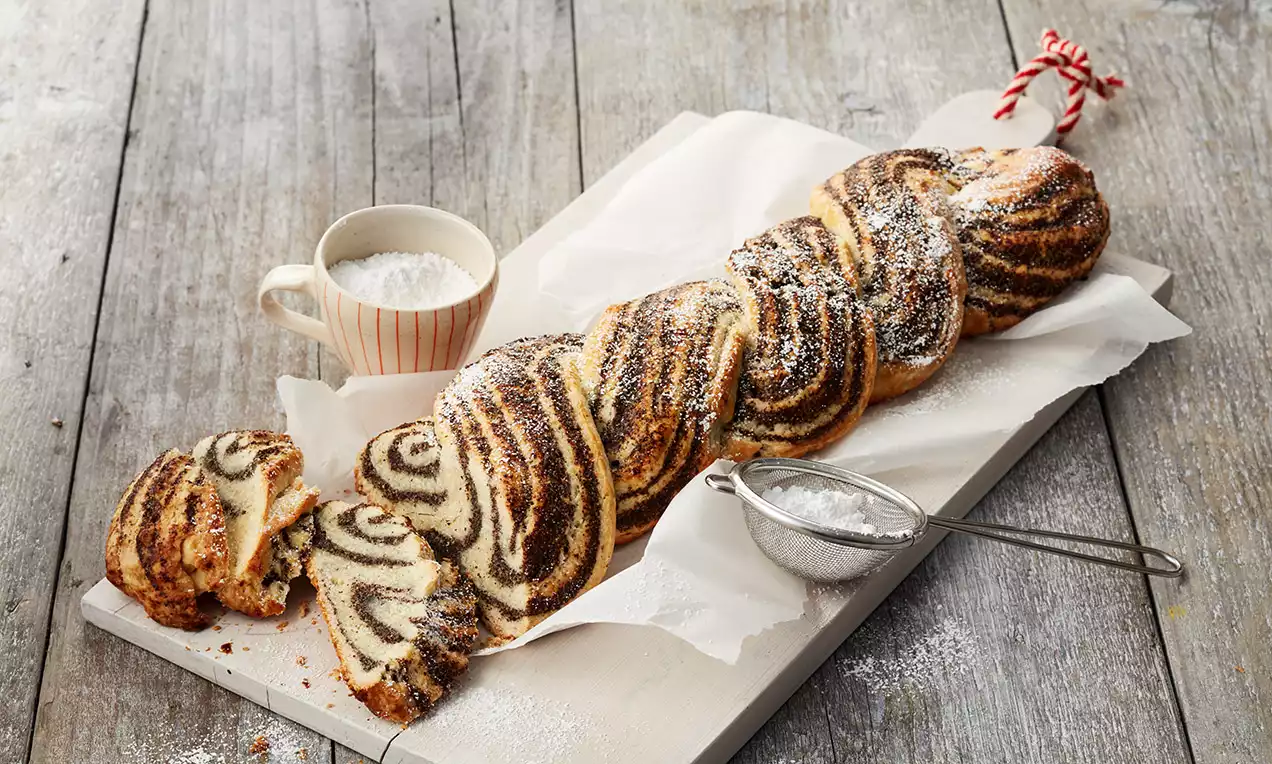 Mohnzopf mit glänzend goldbrauner Kruste und dunkler Mohnfüllung, angeschnitten und mit Puderzucker bestreut auf Holzbrett