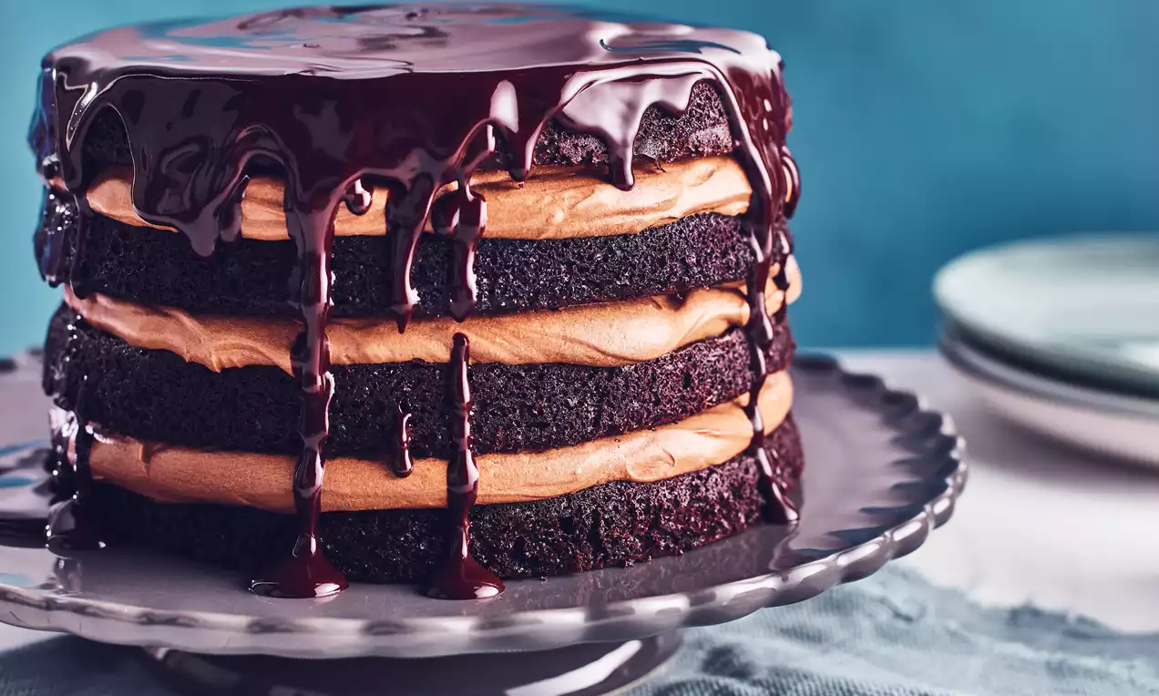 Rich dark chocolate layer cake stacked with smooth caramel-coloured frosting and topped with glossy chocolate ganache on a grey stand