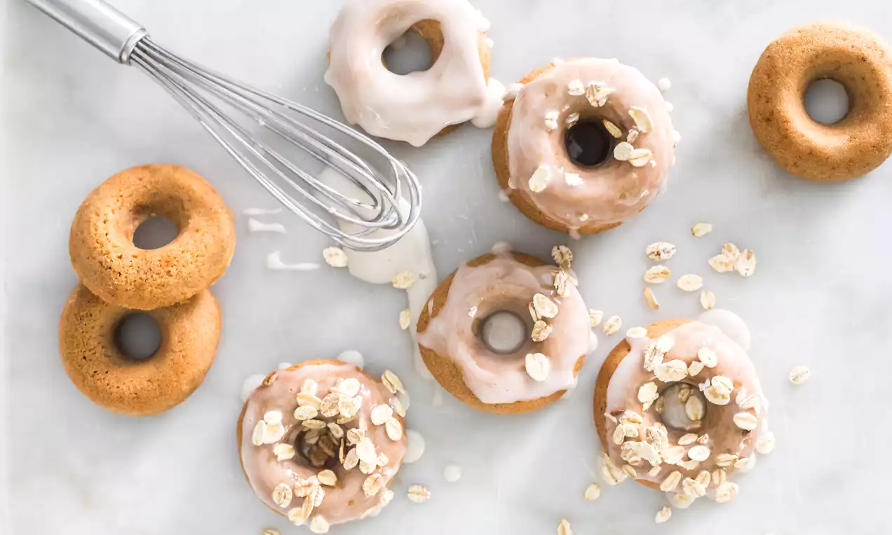 Rosquillas doradas y suaves con glaseado brillante, decoradas con copos de avena y dispuestas sobre mármol blanco junto a un batidor