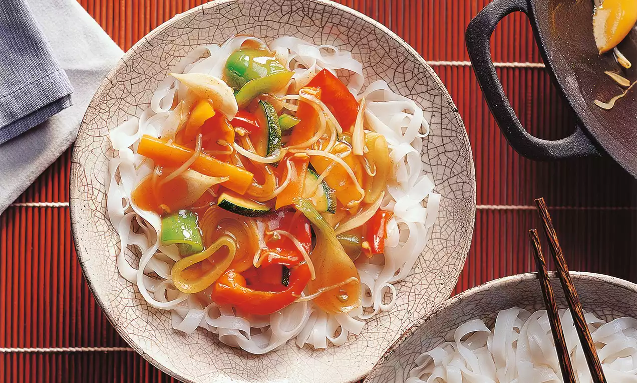 Farebné kúsky zeleniny v lesklej omáčke usporiadané na svetlých ryžových rezancoch na krémovom tanieri s jemným vzorom