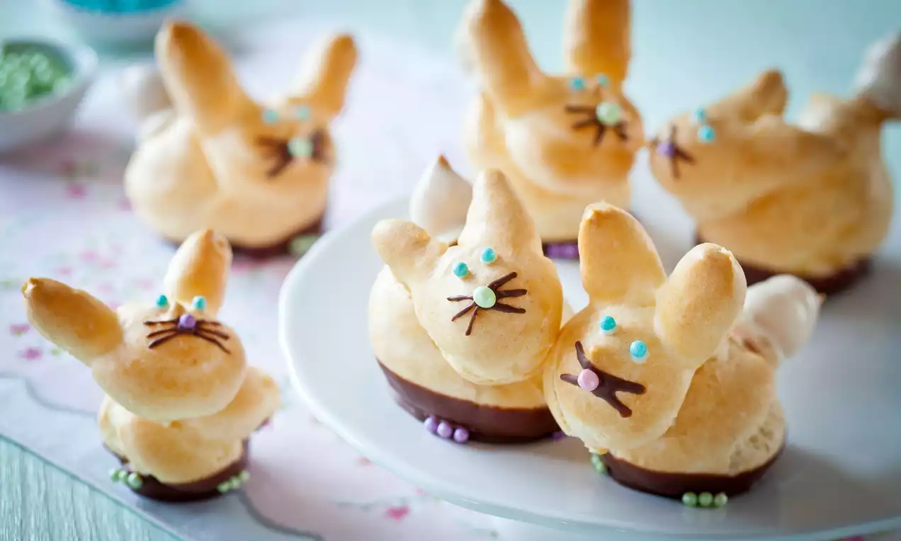 Złociste kruche króliczki z ciasta ptysiowego, dekorowane kolorowymi cukrowymi perełkami, na białym talerzu i pastelowym obrusie