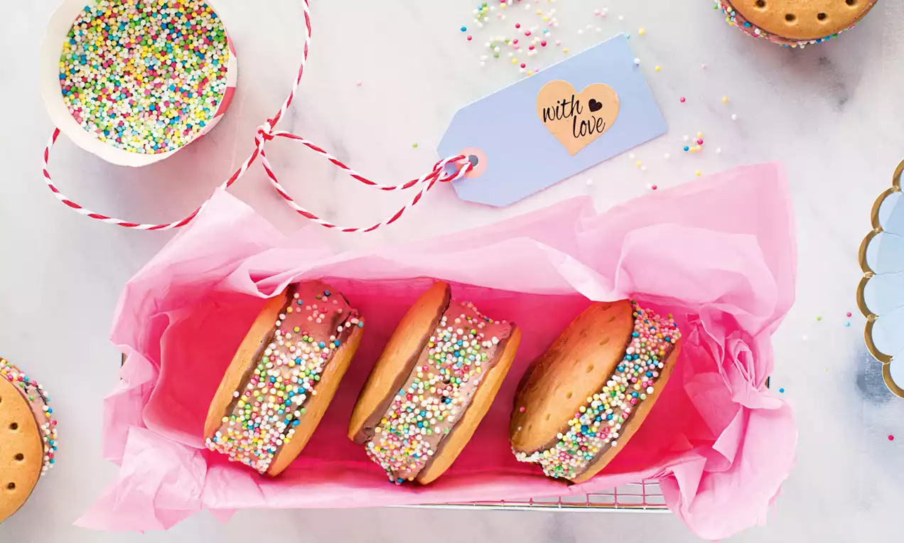 Tri okrogle piškote z rjavimi robovi in vanilijevim čokoladnim sladoledom, okrašene z barvnimi posipi, zavite v rožnat papir