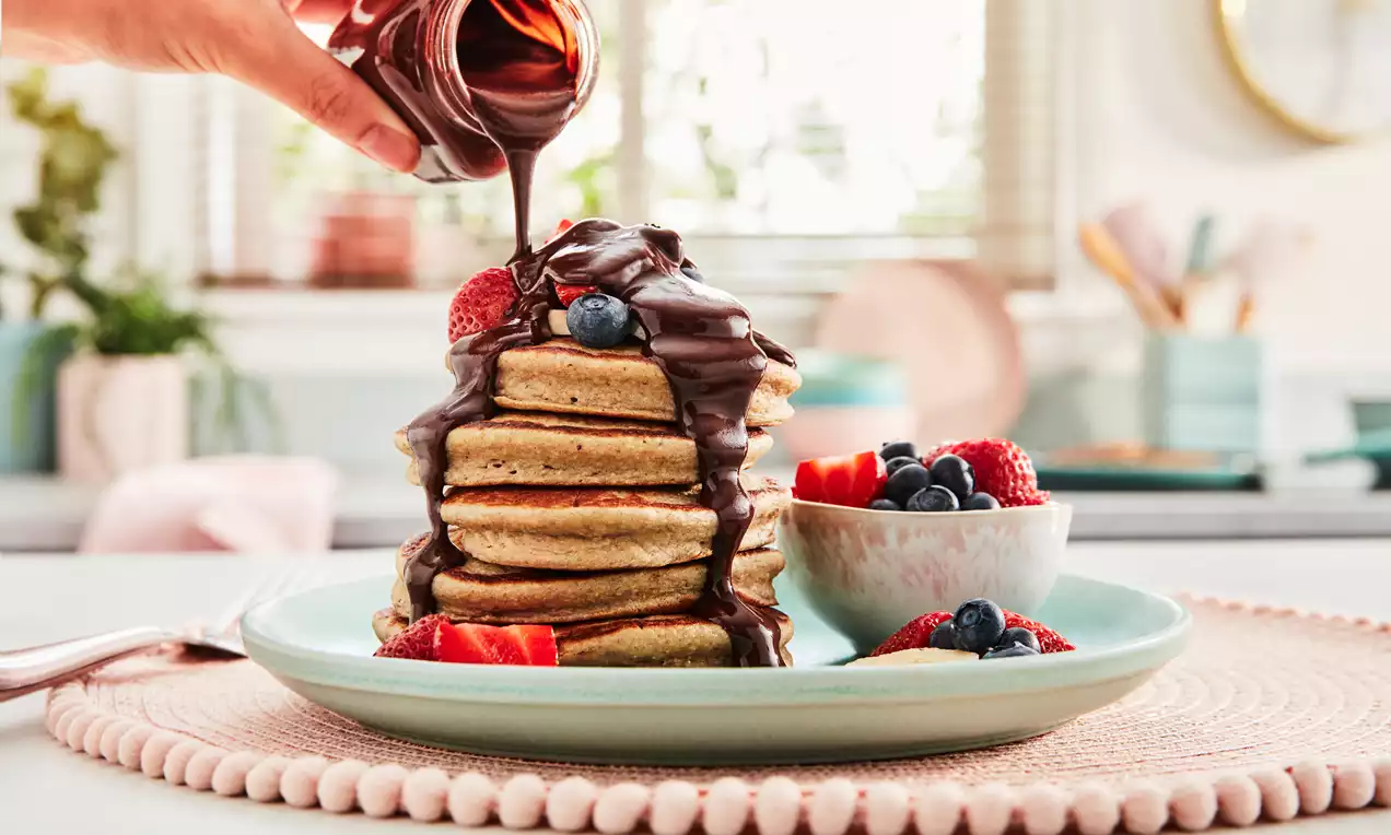 Golden banana pancakes stacked high, drizzled with glossy chocolate sauce, topped with strawberries and blueberries on a pale green plate