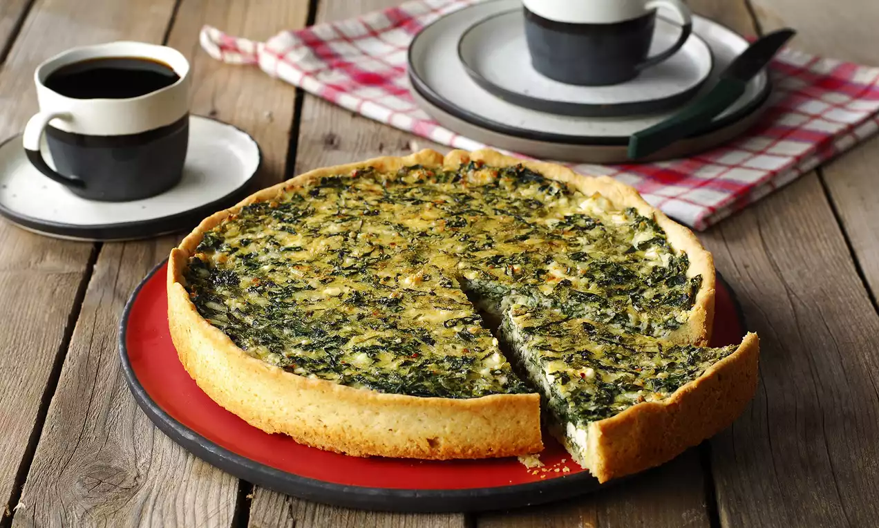 Altın rengi, kıtır tabanlı ıspanaklı kiş, ince bir dilim çıkarılmış, kırmızı tabakta rustik ahşap masada kahve fincanlarıyla sunulmuş