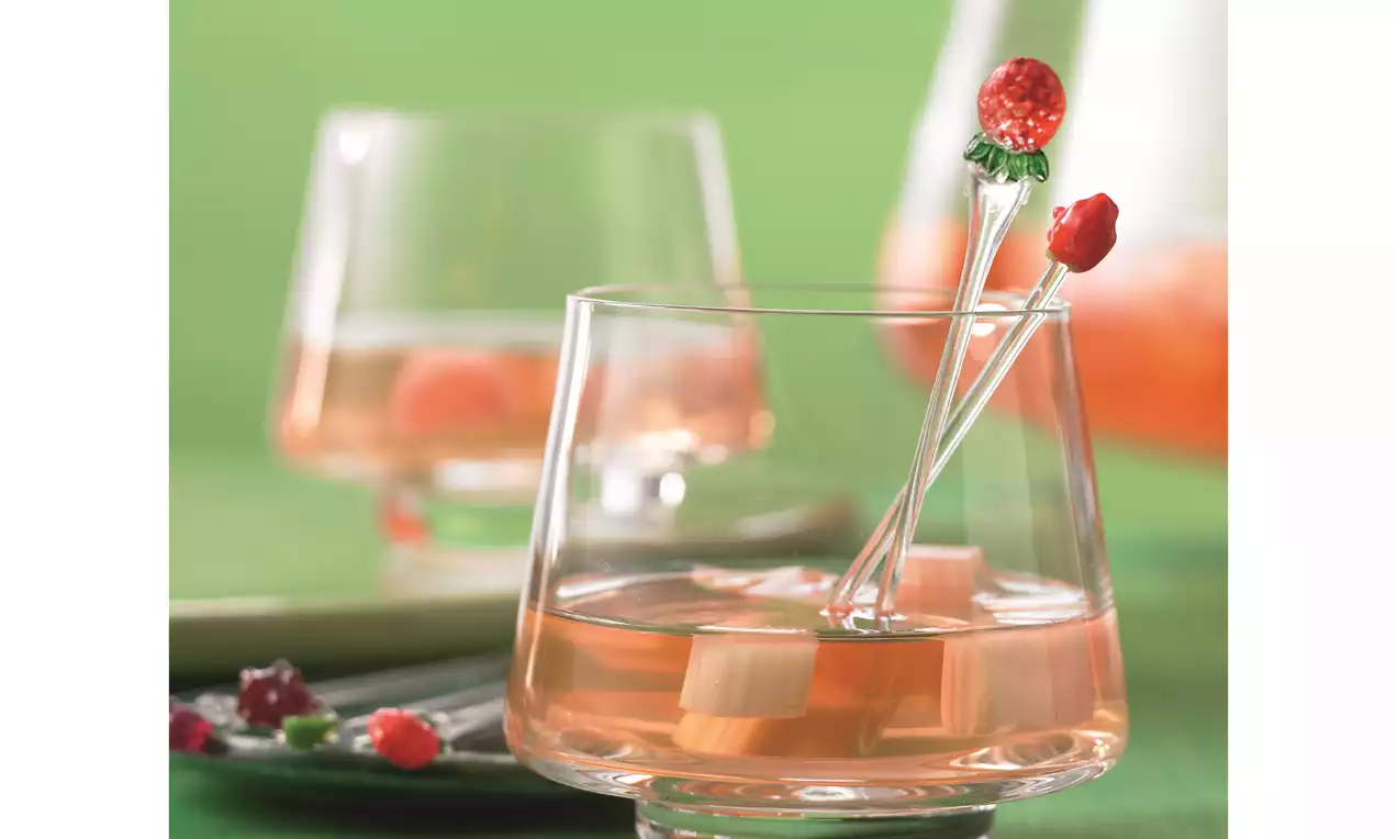 Roséfarbene Rhabarber-Bowle mit kleinen Rhabarberstücken in einem transparenten Glas, dekoriert mit Fruchtspiess auf grünem Tuch