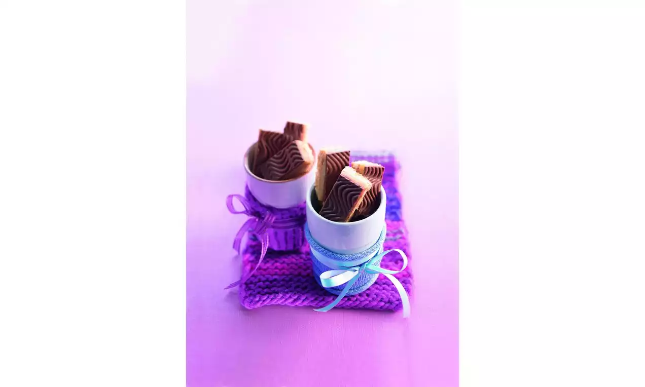 Dva keramické bílé hrnky na fialovém pleteném podkladu, plné karamelových shortbread tyčinek s vlnkovaným vzorem, ozdobené stuhami