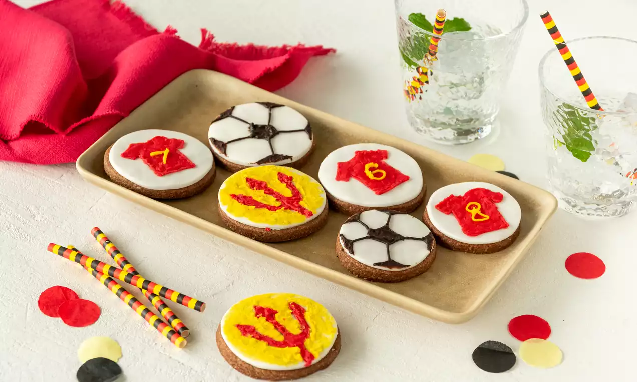 Voetbalkoekjes met witte en gele icing, versierd als ballen, rode shirtjes en een duivel op een beige schaal met feestelijke decoratie