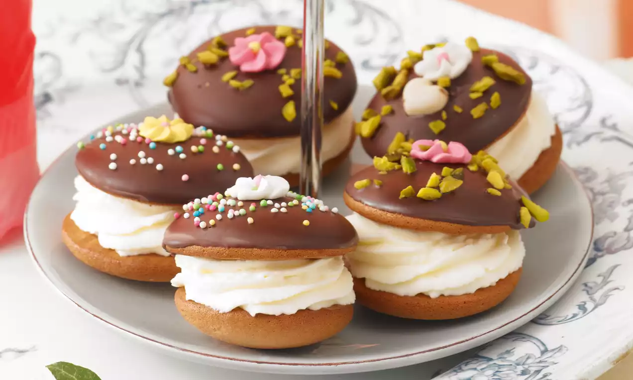 Indianerkrapfen mit goldbraunem Gebäckboden, weißer Sahnefüllung und glänzender Schokoladenglasur, verziert mit Pistazien und Zuckerblüten