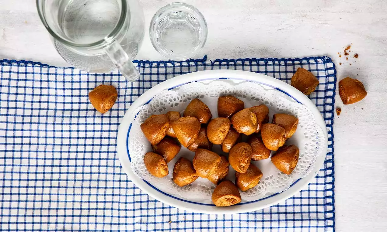 Piccoli dolci dorati dalla forma irregolare, con superficie lucida e porosa, disposti in una ciotola bianca su un panno a quadretti blu