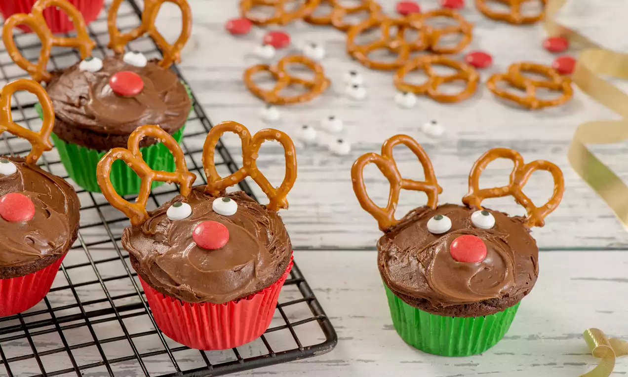 Chocolate cupcakes in red and green liners with pretzel antlers, white sugar eyes, and red candy noses on a cooling rack on a wooden table