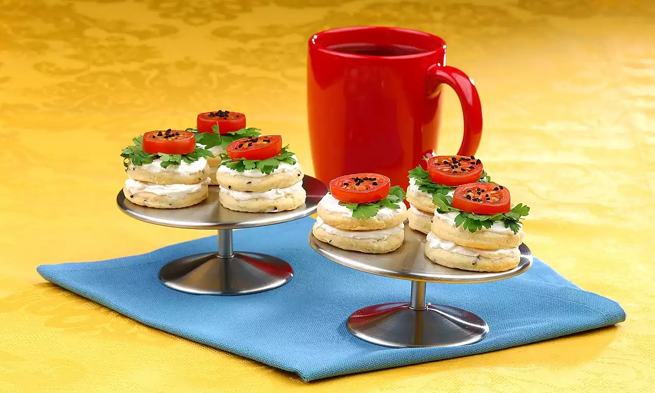 Yuvarlak metal standlarda, labne peynirli, maydanoz yapraklı, domates dilimli ve çörekotlu kanepeler, mavi bir örtü üzerinde sıralanmış