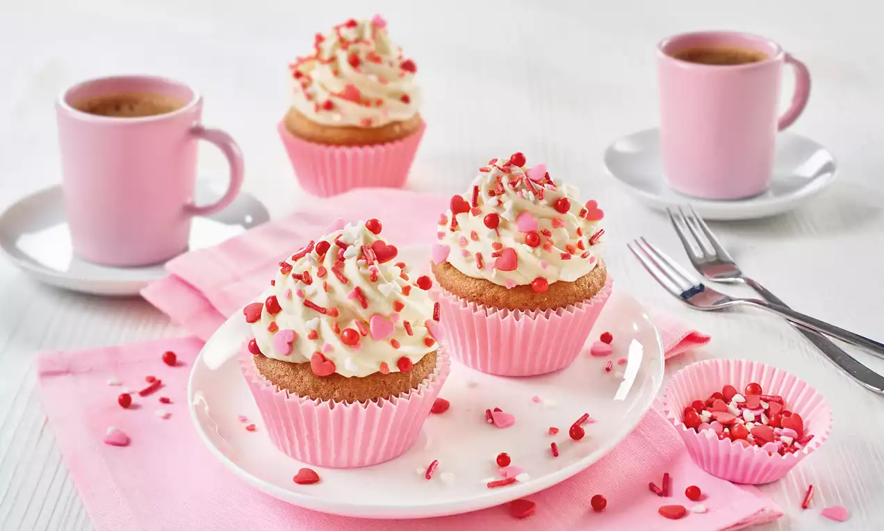 Muffins mit weißer Sahnehaube und roten Herzstreuseln in rosa Papierförmchen auf einem weißen Teller mit rosa Tasse und Besteck