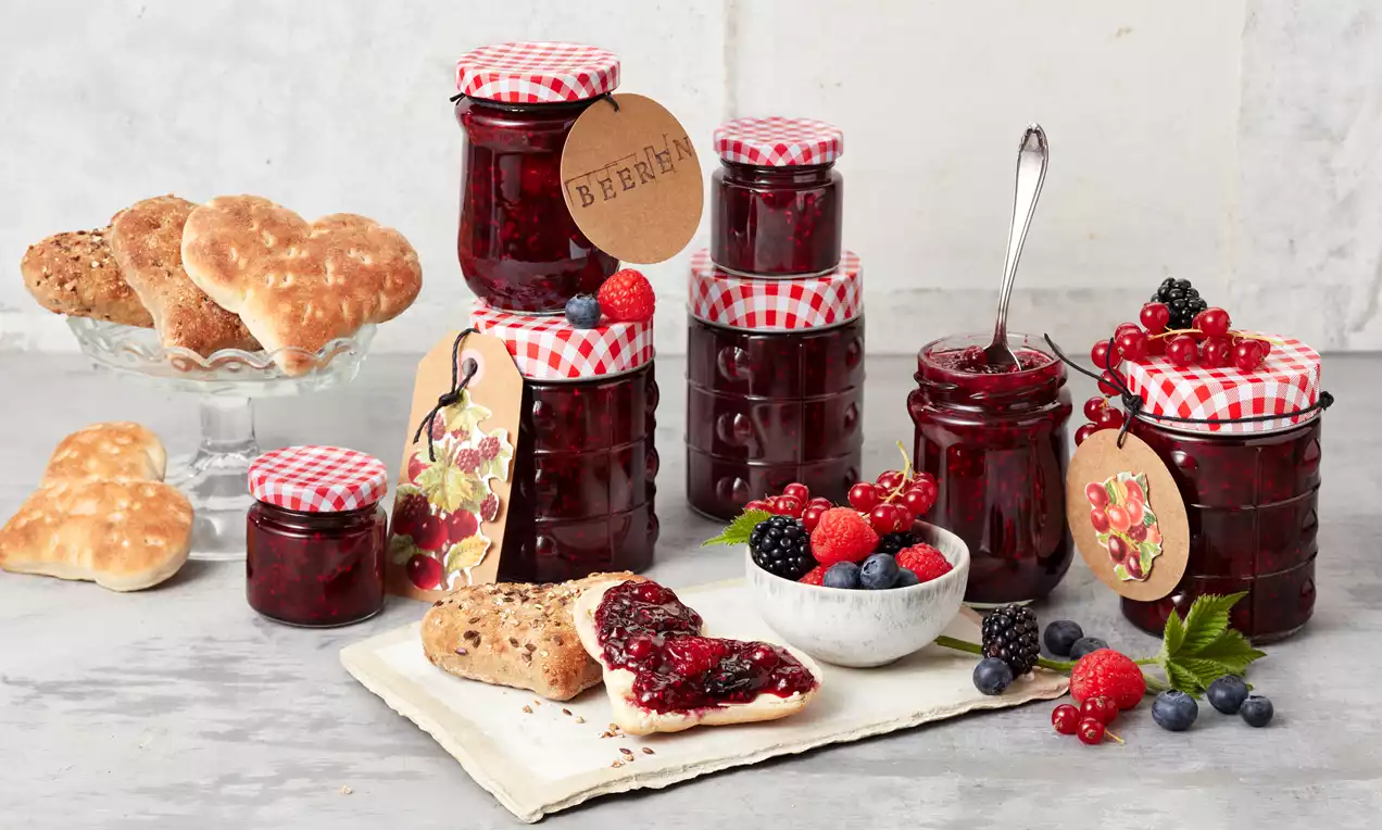 Gläser mit dunkelroter Beerenmarmelade und rot-weiß karierten Deckeln, dekoriert mit Beeren und Herzgebäck auf einer hellen Platte