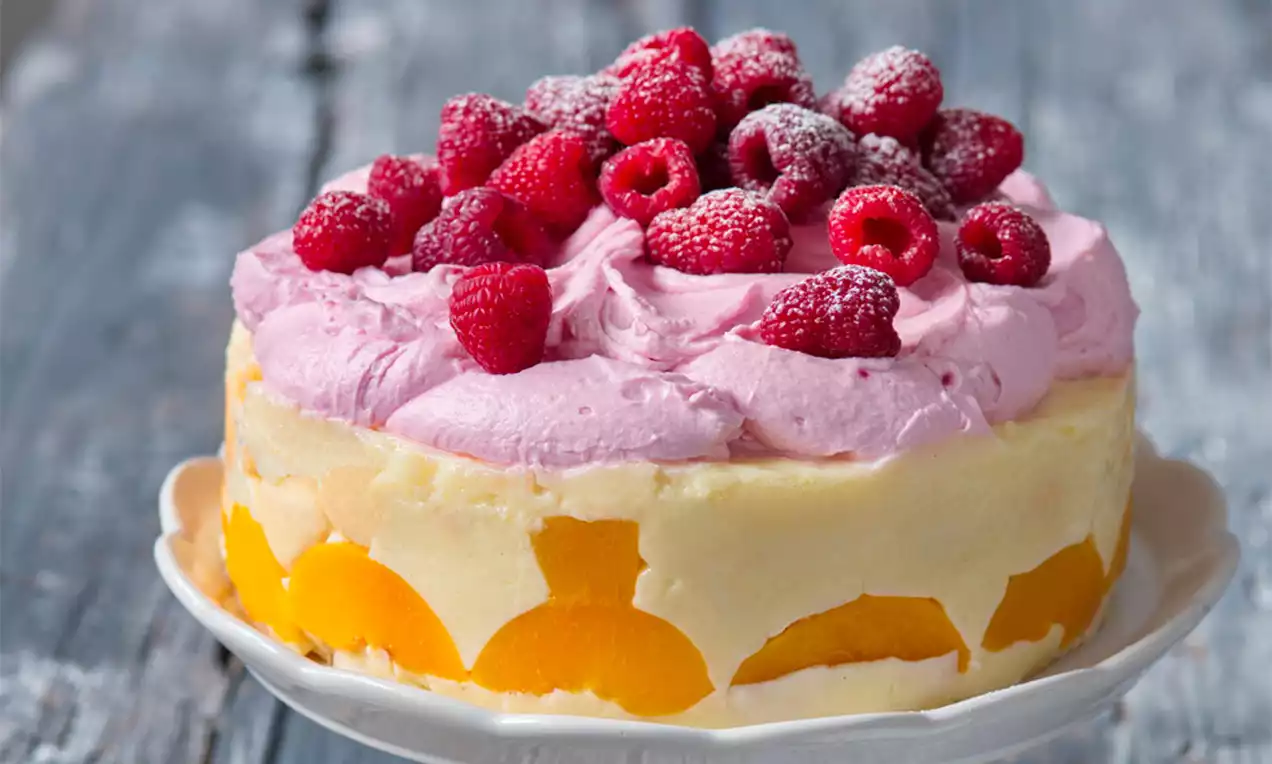 Pudingový dort s vrstvou žlutých broskví, jemnou vanilkovou náplní, růžovou šlehačkou a malinami posypanými moučkovým cukrem na bílém talíři