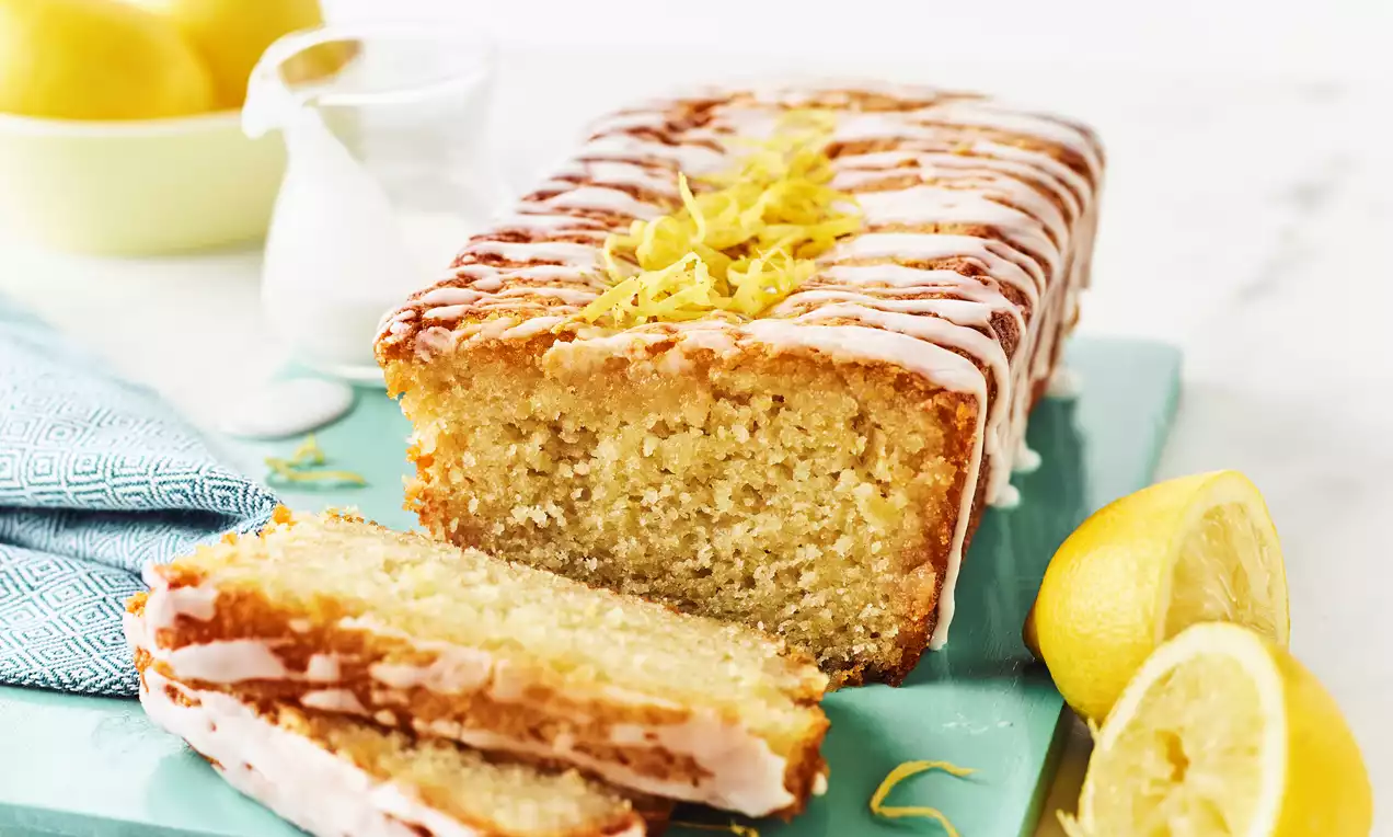 Golden vegan lemon drizzle loaf with moist crumb, topped with white icing and lemon zest, sliced on a pastel blue board surrounded by fresh lemons