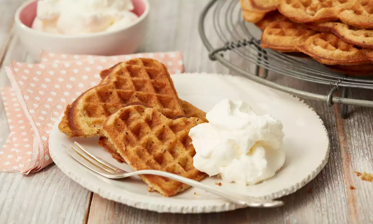 Goldbraune, herzförmige Nuss-Waffeln mit luftigem Eisberg Sahne auf einem weißen Teller neben rosa Stoffserviette und Gabel