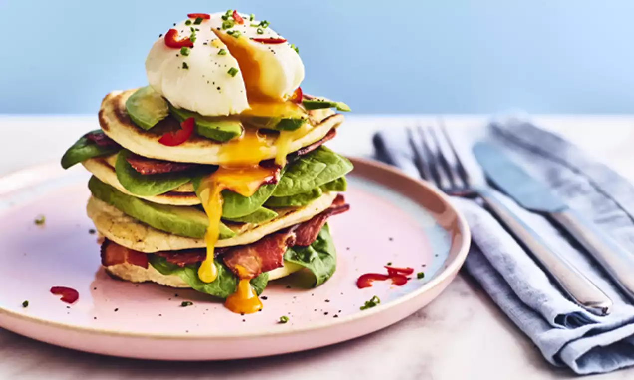 A stack of golden pancakes layered with green avocado slices, crispy bacon, a runny poached egg, and red chilli slices on a pink plate