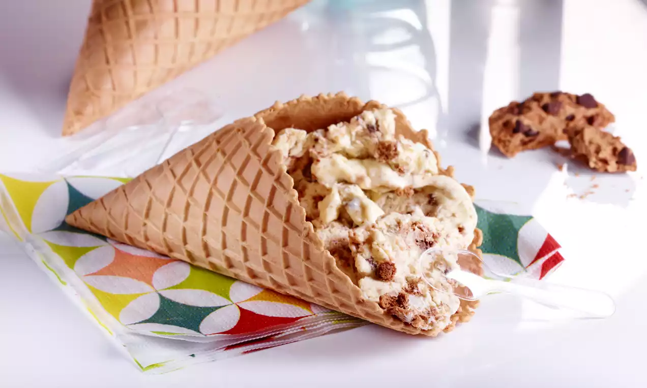 Knusprige Waffel mit cremigem Eis voller brauner Chocolate-Chip-Stücke auf einem bunten Serviettendruck, daneben ein Stück Keks
