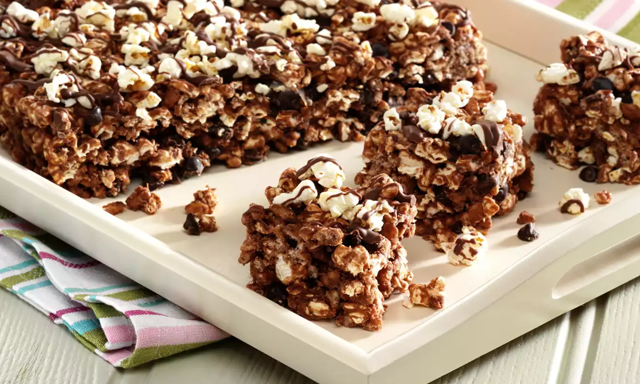 Squares of chocolate-coated cereal and popcorn clusters drizzled with dark chocolate on a cream tray with a striped cloth underneath