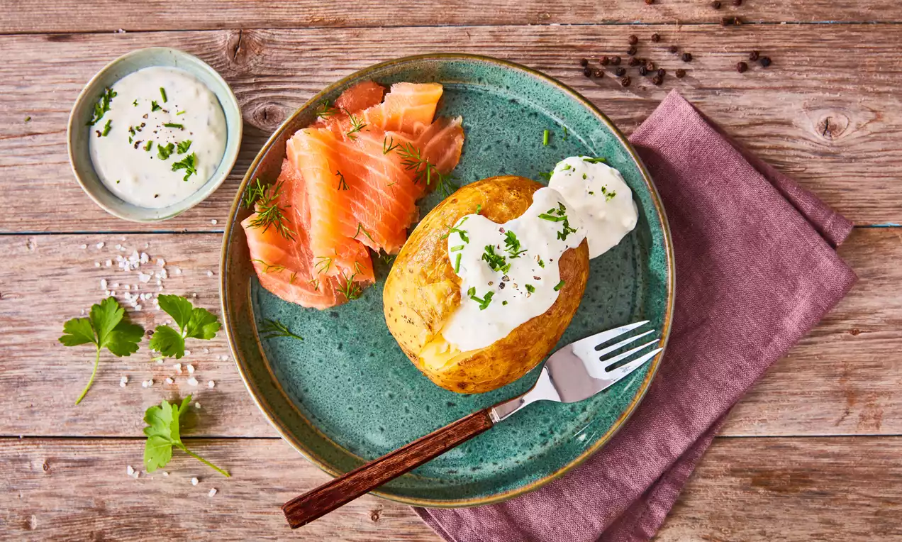 Goldgelbe Folienkartoffel mit weißer Sour Cream und grünen Kräutern neben zartem Lachs auf türkisfarbenem Teller mit Holzuntergrund