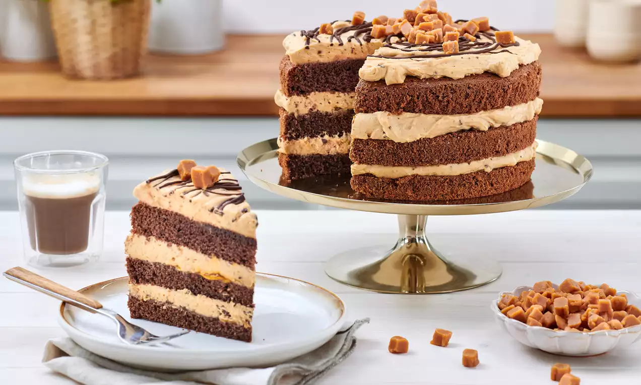 Triple-layer chocolate caramel cake with caramel cream frosting, drizzled chocolate, and caramel cubes on a golden cake stand