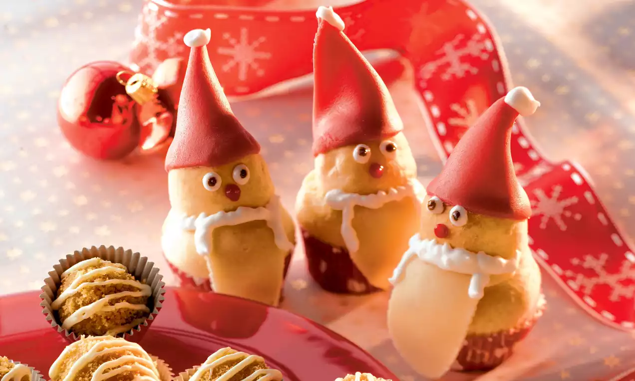 Kırmızı şapkalı kremalı mini kekler, beyaz süslemelerle detaylandırılmış, kırmızı tabakta kurabiye yanında sunulmuş, arka planda kırmızı kurdele