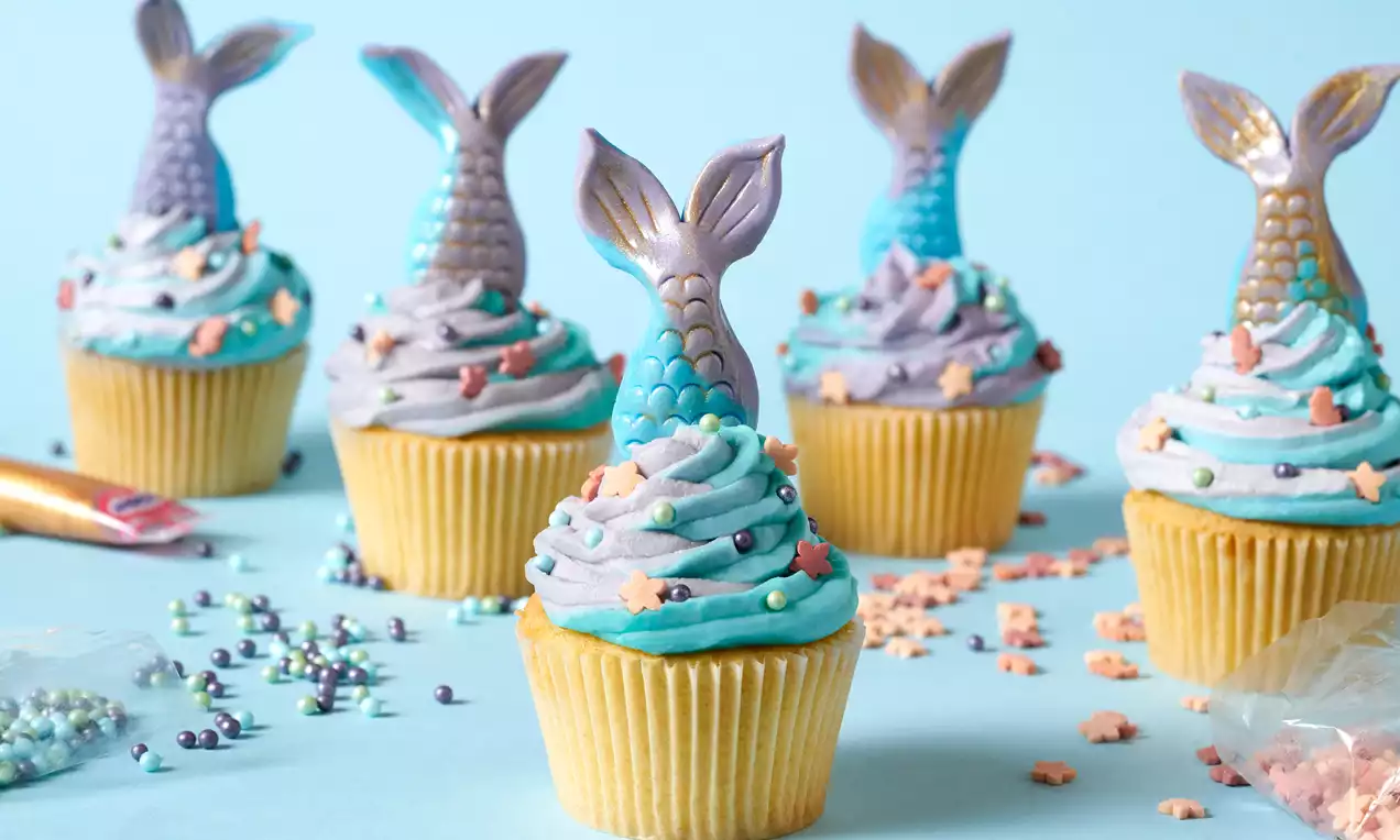 Cupcakes with swirls of blue and lilac frosting, topped with shimmering mermaid tails and scattered with pearl and star sprinkles