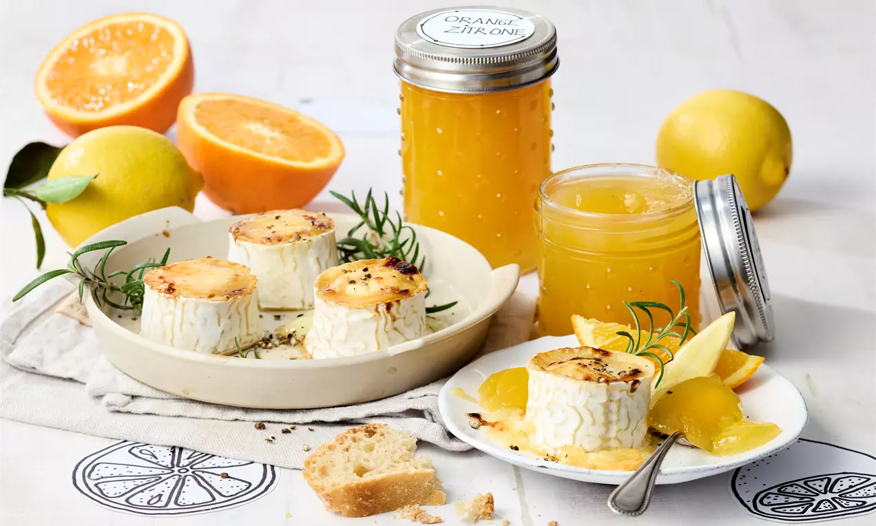 Orangen-Zitronen-Gelee in offenen Gläsern, dazu karamellisierter Käse auf weißem Teller mit Rosmarin, Zitrusscheiben und Brotstücken drapiert