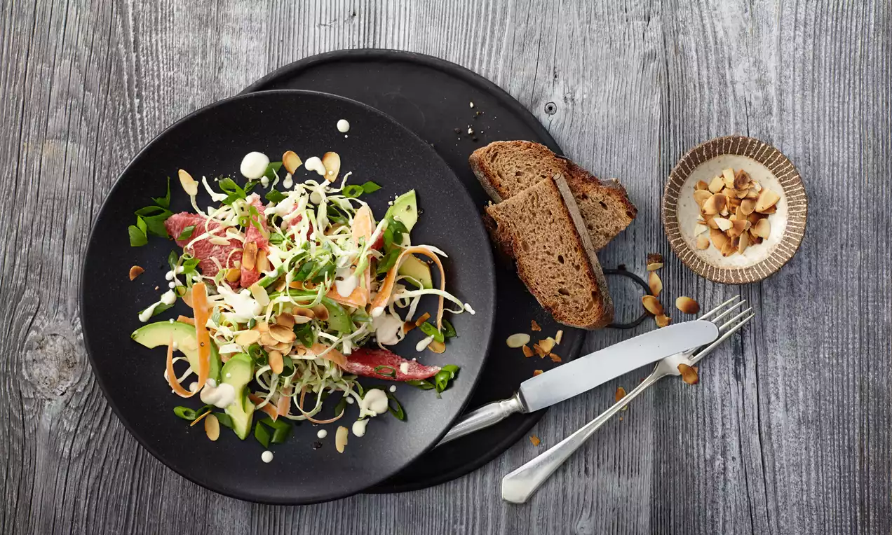 Feiner Spitzkohlsalat mit dünnen Karottenstreifen, Gurkenstücken, Kresse und Dressing auf schwarzem Teller, serviert mit Brotscheiben und Nüssen