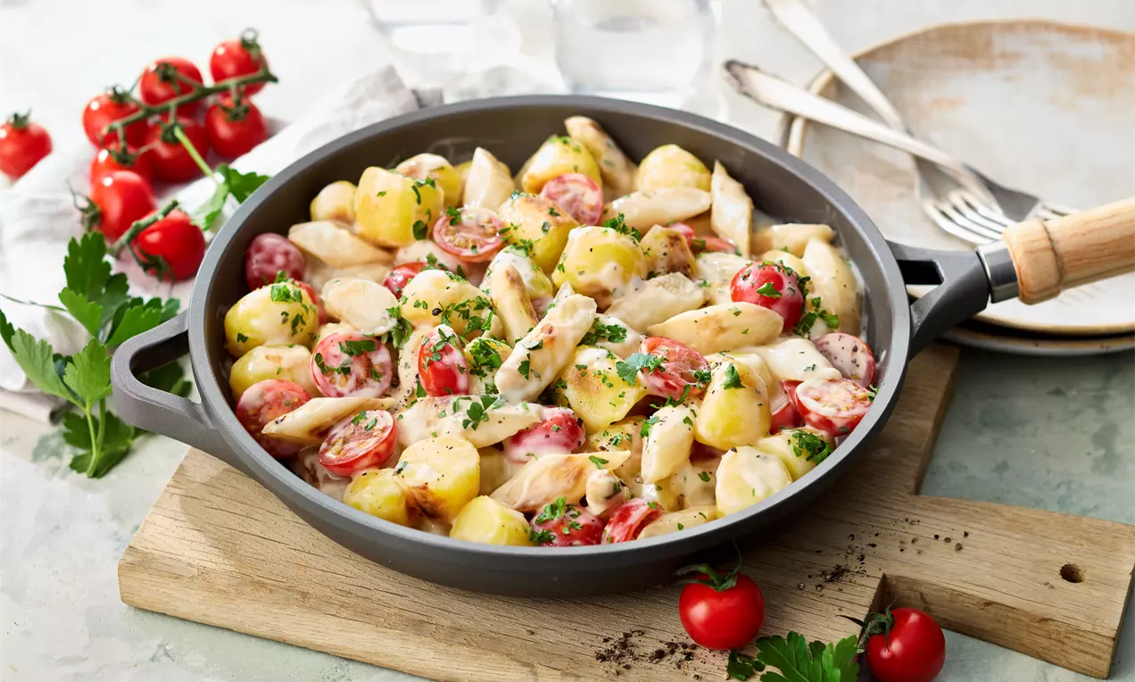 Cremige Spargelpfanne mit weißen Spargelstücken, gelben Kartoffelwürfeln, roten Tomatenhälften und frischen Kräutern in einer grauen Pfanne