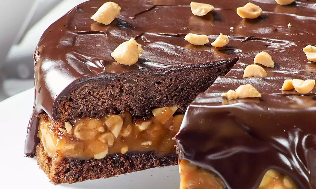 Čokoladni kolač s tamnim sjajnim preljevom, slojem kikiriki karamele i posutim cijelim komadićima kikirikija na bijelom tanjuru