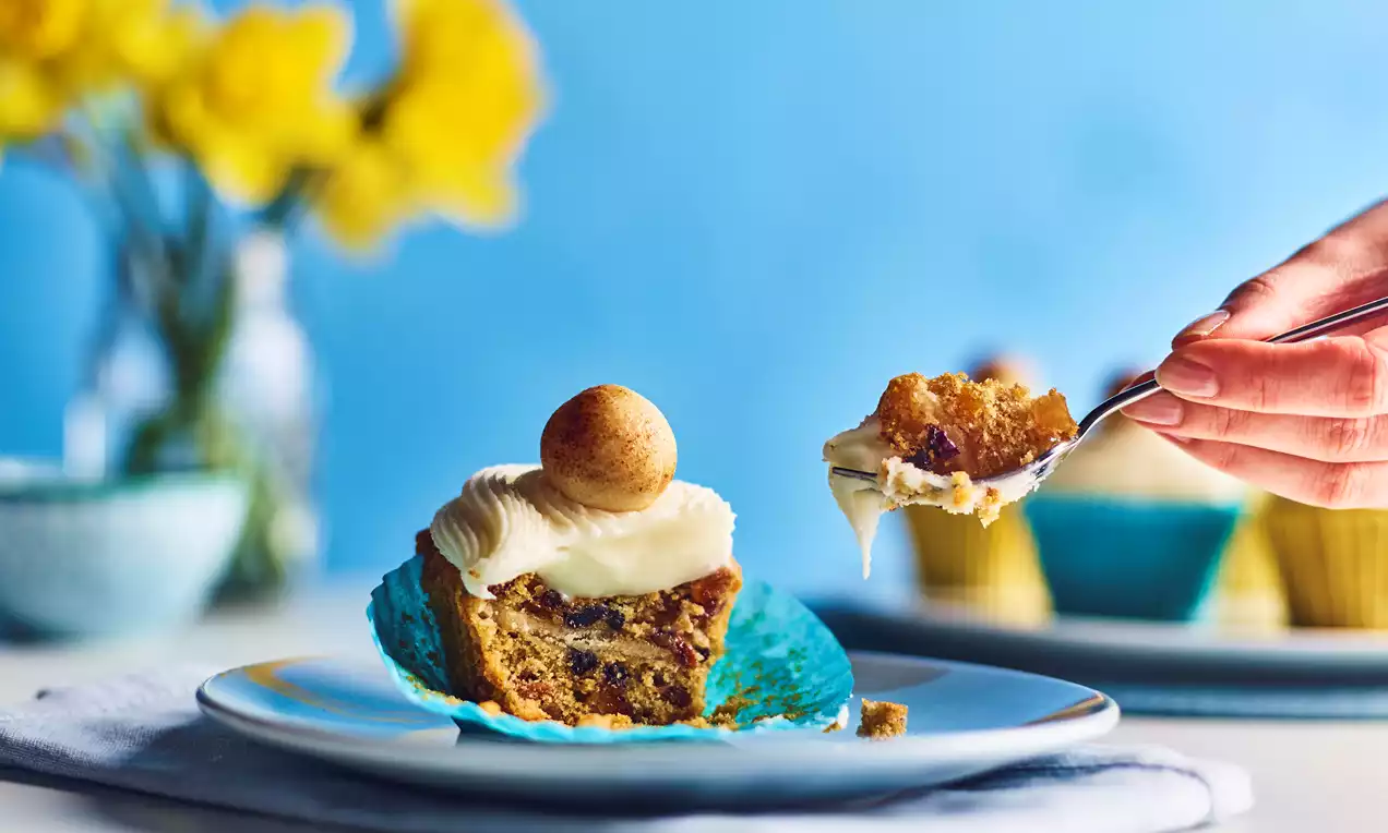 A Simnel cupcake with golden crumb dotted with fruit, topped with white cream and an almond ball, set on a plate with a blue wrapper